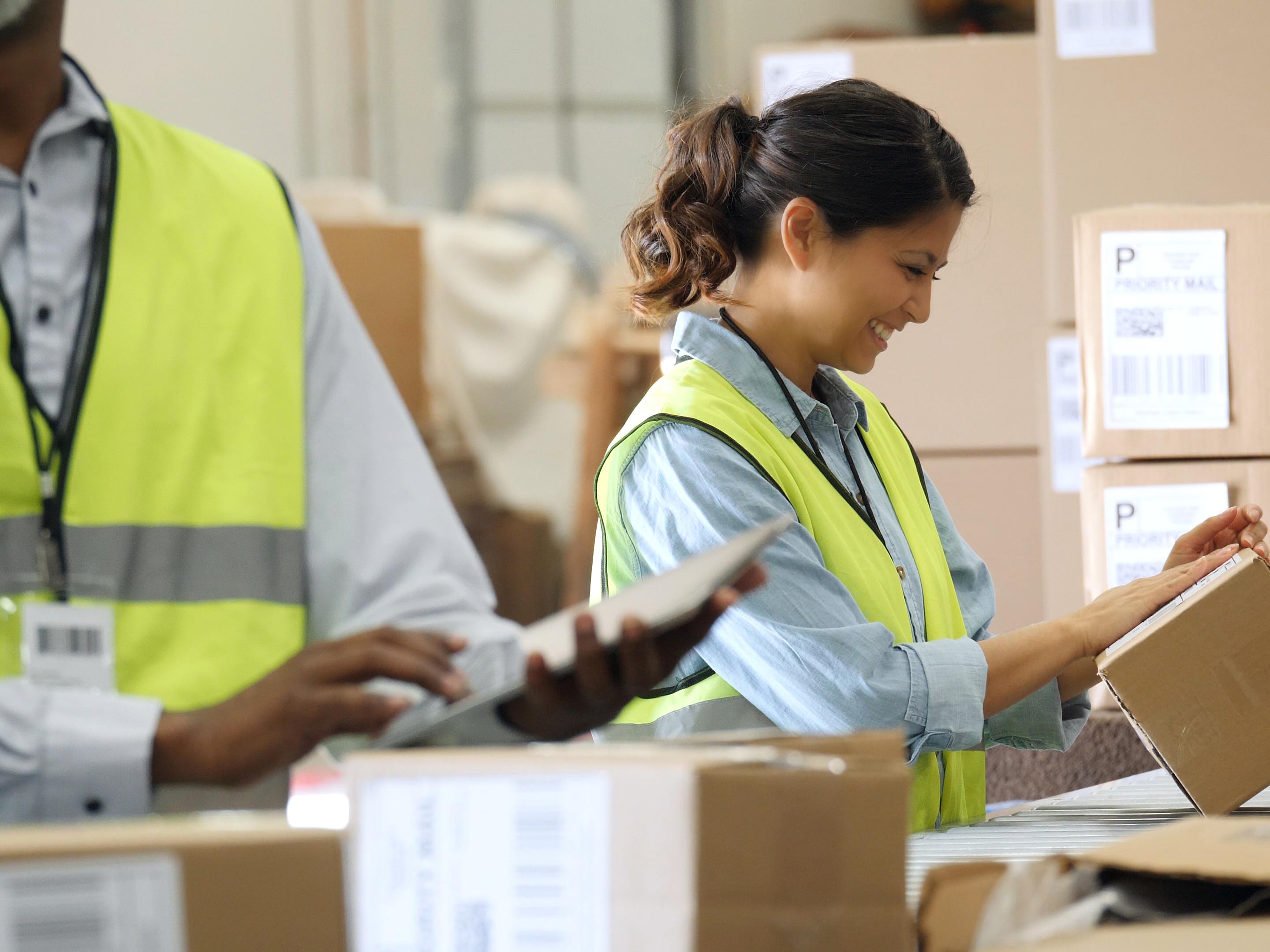Confident male and female distribution warehouse workers prepare to send out packages to customers. A female employee is placing a mailing label on the package. A male employee is using a digital table tin the foreground.
