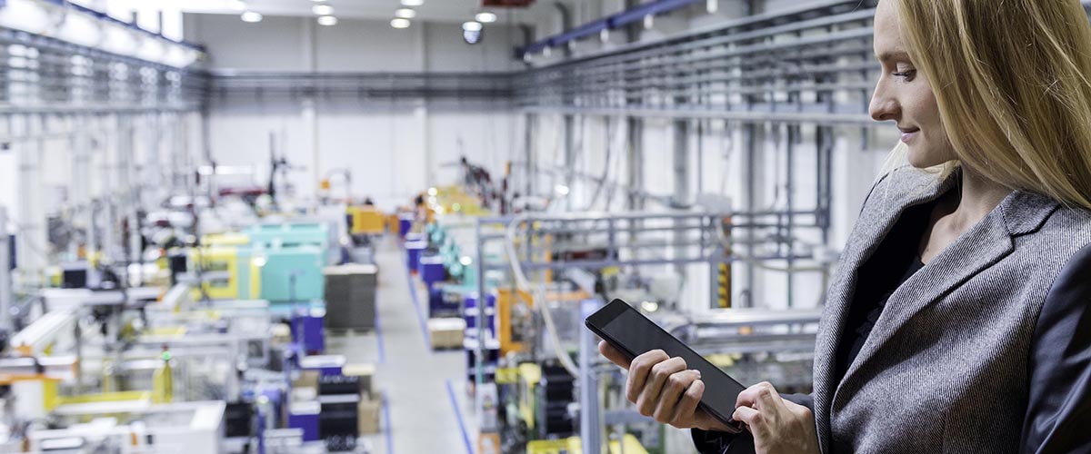 Horizontal color image of blond manager working with digital tablet in large futuristic factory. Blond woman standing on top of a balcony. Focus on businesswoman, futuristic machines in background.