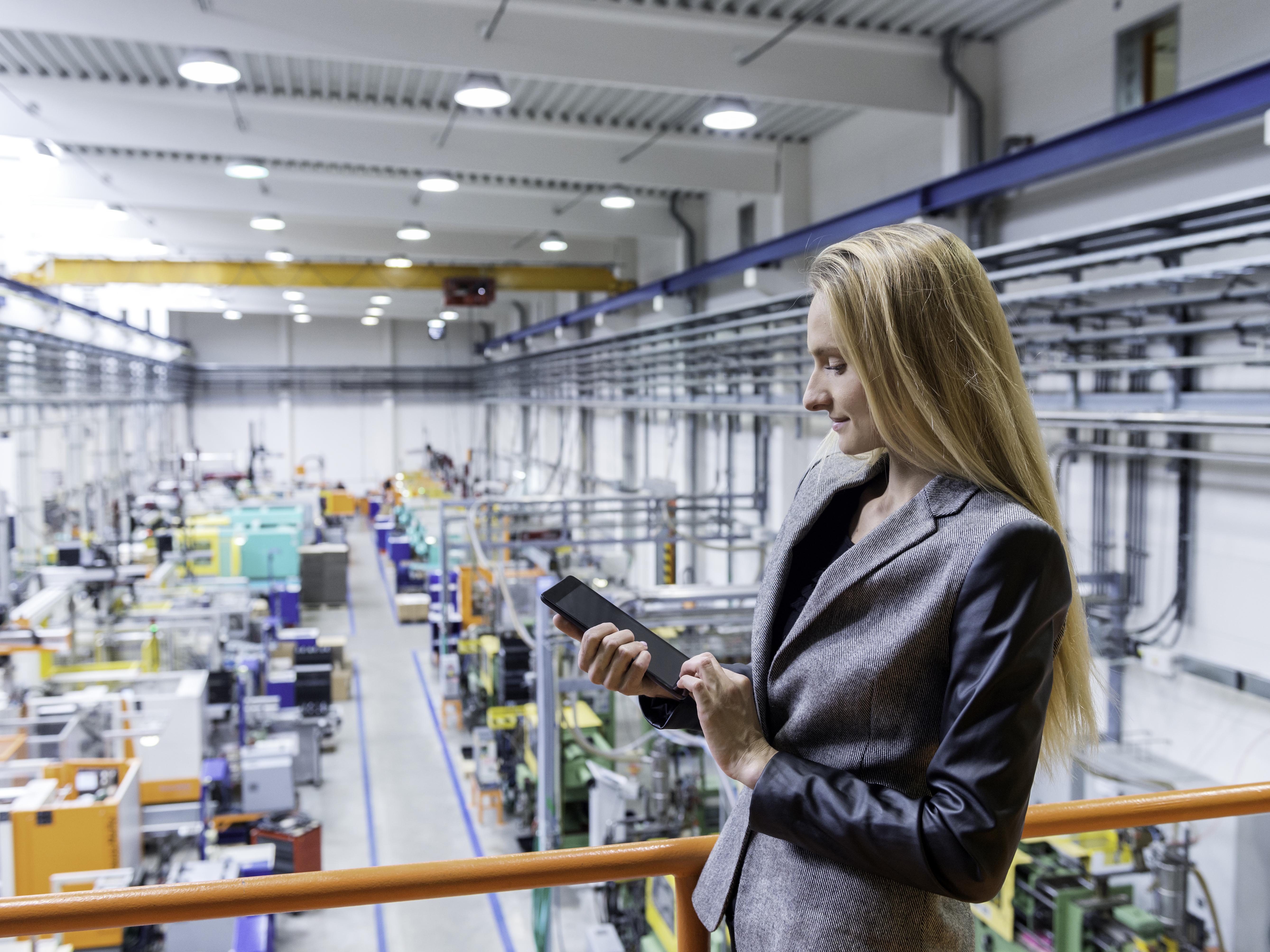 Horizontal color image of blond manager working with digital tablet in large futuristic factory. Blond woman standing on top of a balcony. Focus on businesswoman, futuristic machines in background.