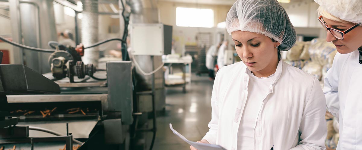 Two quality professionals in white sterile uniforms checking quality of salt sticks while standing in food factory.