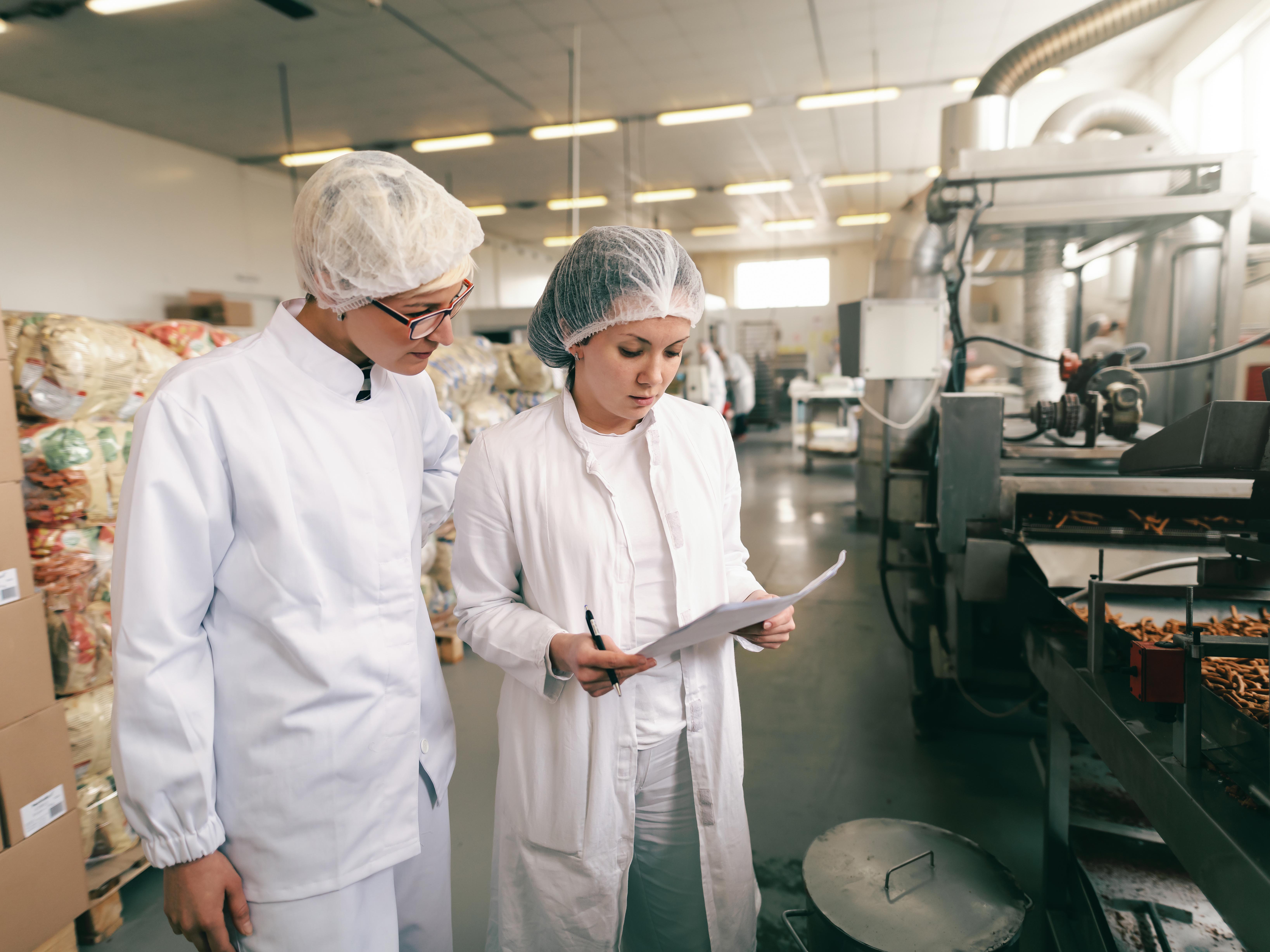 Two quality professionals in white sterile uniforms checking quality of salt sticks while standing in food factory.